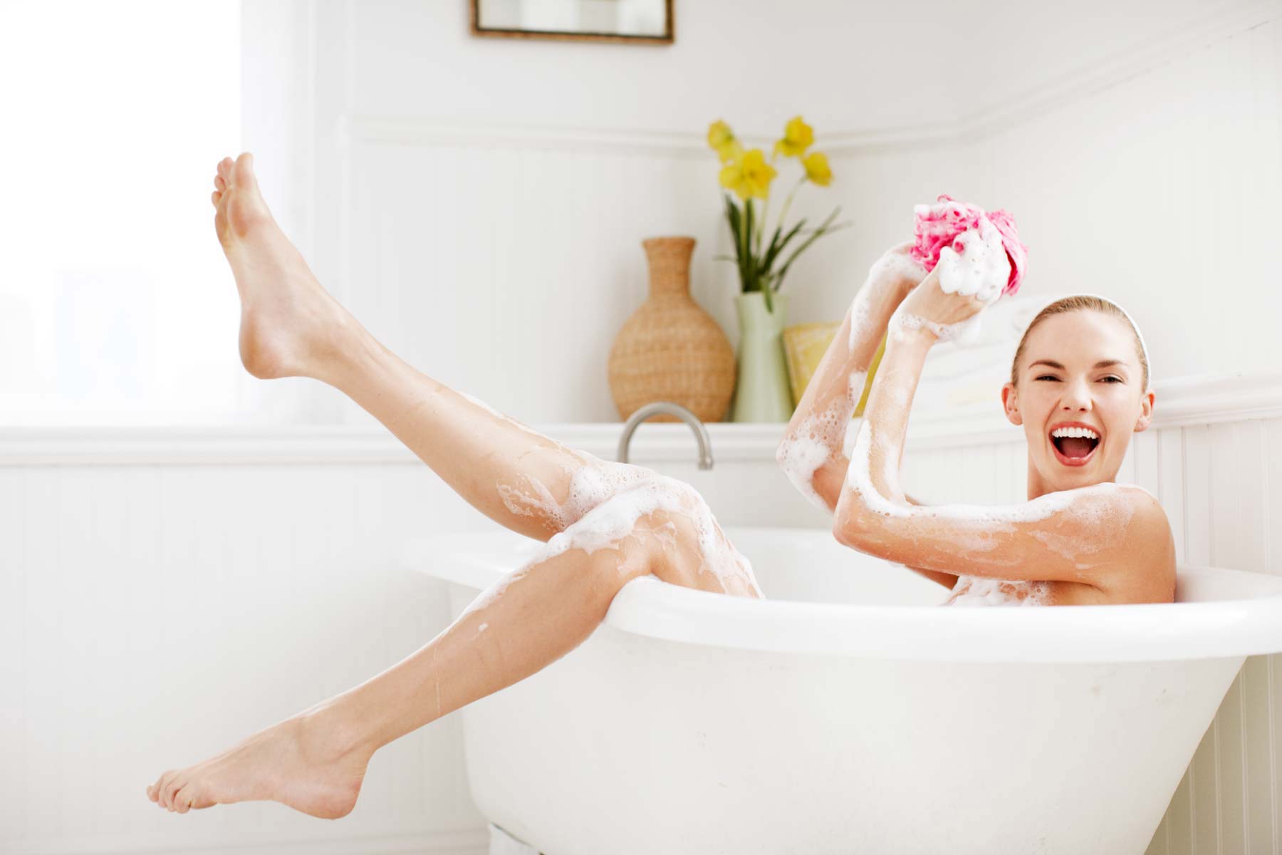Smiling woman relaxing in a bubble bath, legs raised, holding pink sponge in a bright bathroom. Self-care and wellness concept.