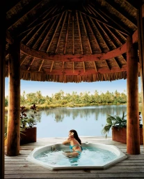 Woman relaxing in an outdoor hot tub under a thatched wooden pavilion overlooking a calm lake and distant trees.
