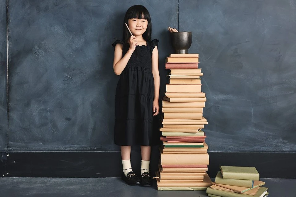 A young girl in a black dress stands next to a tall stack of books in front of a dark blue textured wall, holding a pencil.