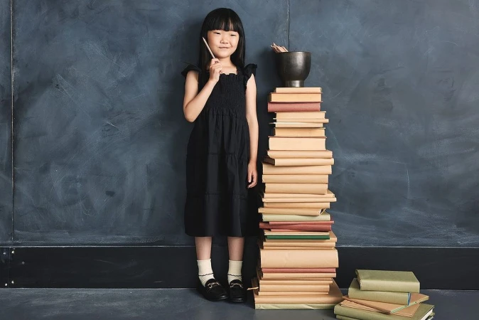 A young girl in a black dress stands next to a tall stack of books in front of a dark blue textured wall, holding a pencil.