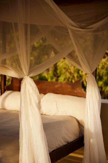 Bedroom with a wooden four-poster bed draped with white curtains, pillows, and bedding, with an outdoor view through the bedroom window.