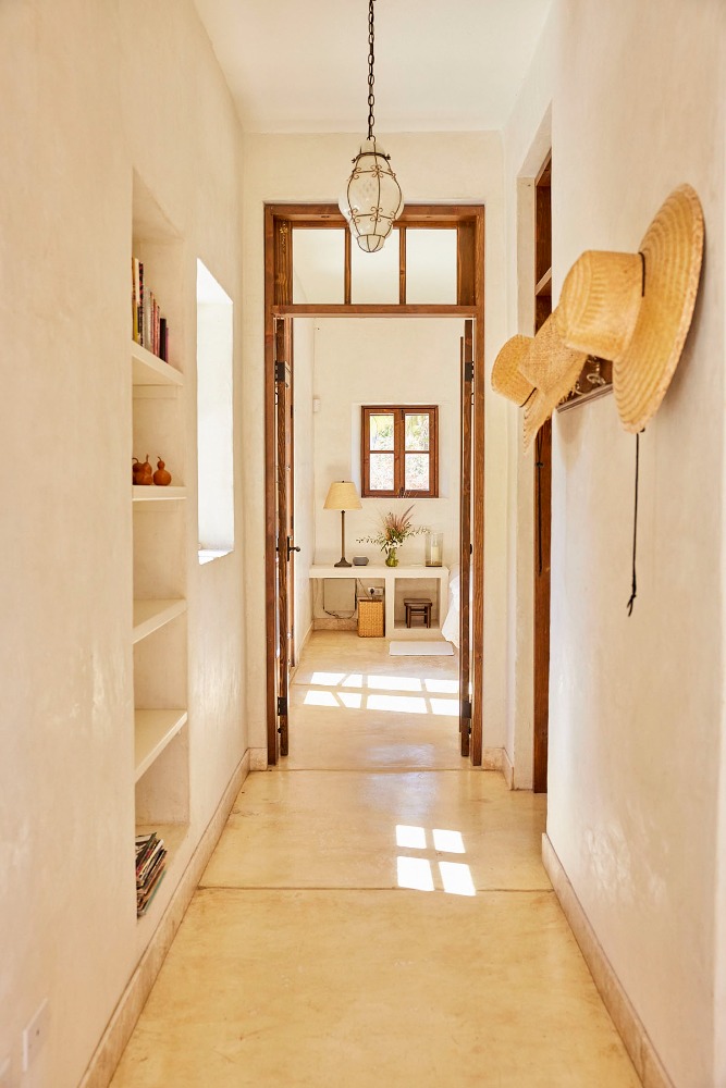 Interior view of a bright hallway with straw hats on the wall, leading to a room with a table and window.