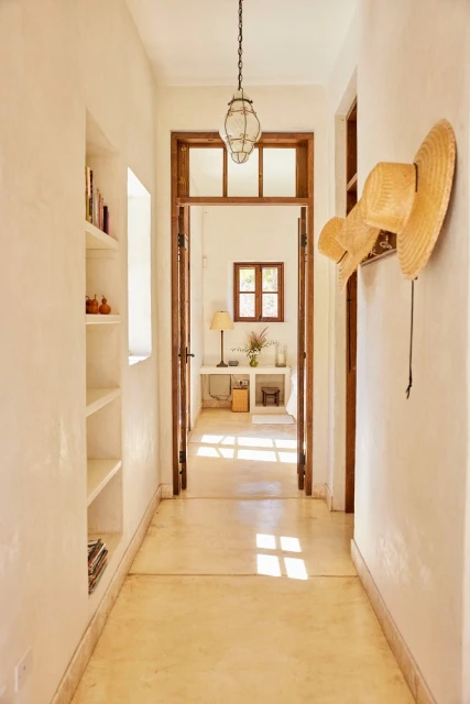 Interior view of a bright hallway with straw hats on the wall, leading to a room with a table and window.
