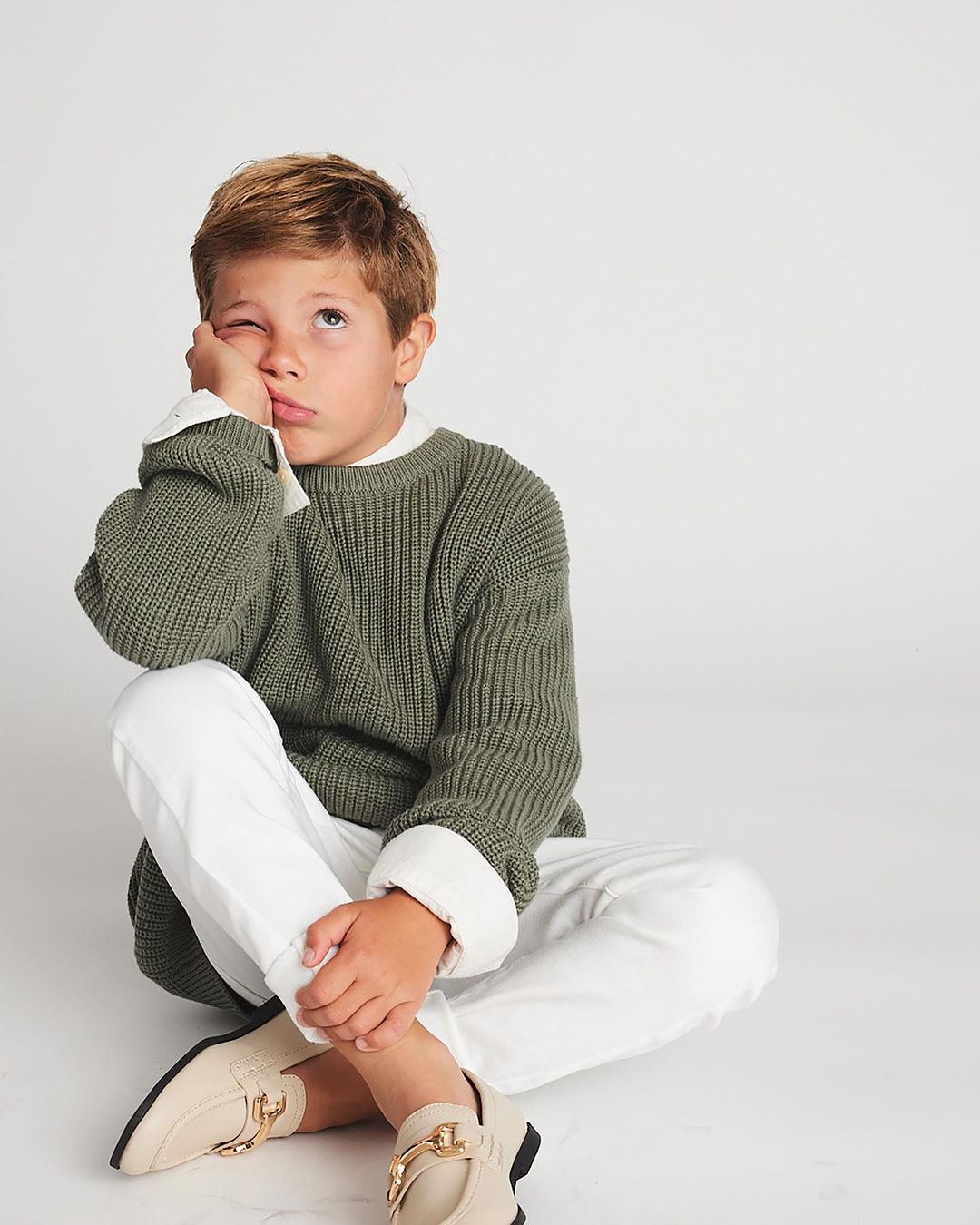 Boy with blond hair sitting cross-legged in a photo studio wearing a green sweater, white pants, and beige moccasins with gold hardware.