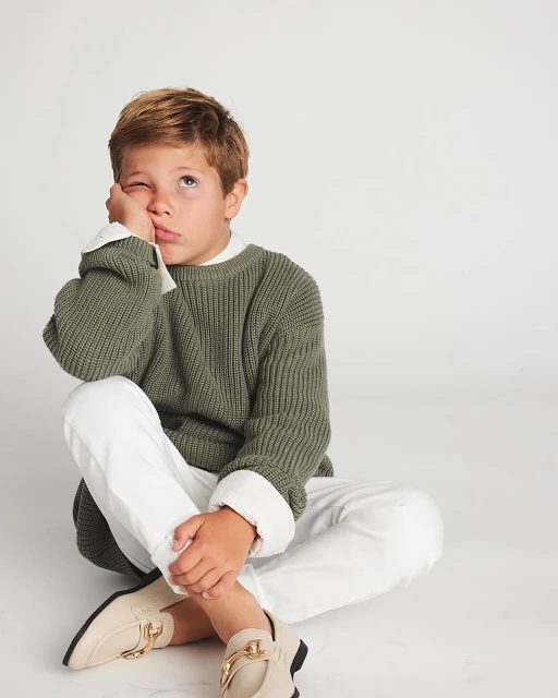 Boy with blond hair sitting cross-legged in a photo studio wearing a green sweater, white pants, and beige moccasins with gold hardware.