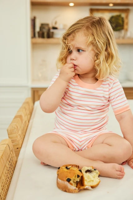 Child with blonde curly hair sitting with crossed legs next to a muffin on a white counter in a kitchen, looking thoughtfully to the side.