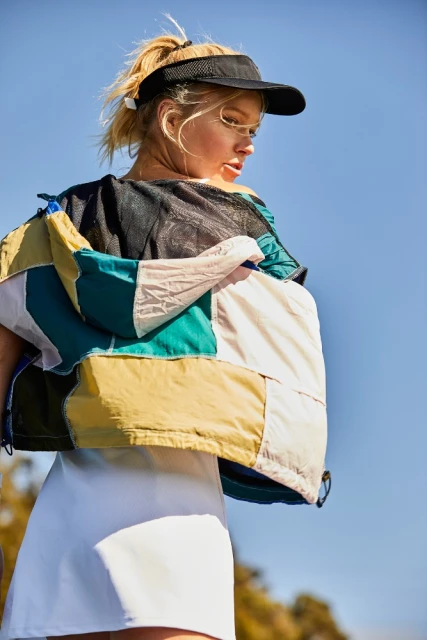 Blonde woman wearing a black visor and a white dress posing outdoors against a blue sky, with a colorful patchwork jacket.