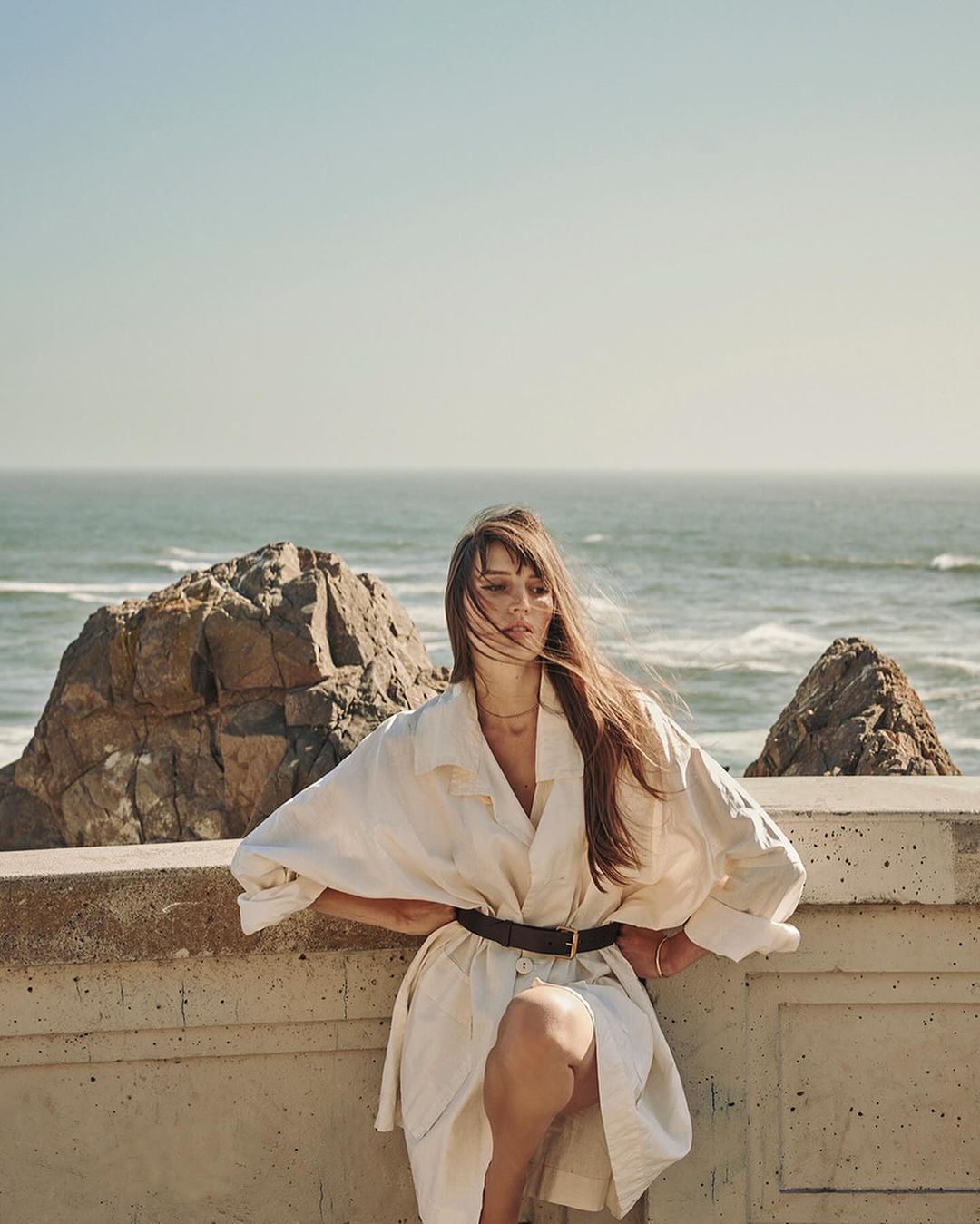 Woman in a white trench coat poses on a concrete wall near the ocean with rocks on the beach. Clear blue sky.