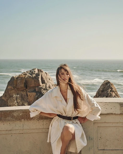 Woman in a white trench coat poses on a concrete wall near the ocean with rocks on the beach. Clear blue sky.