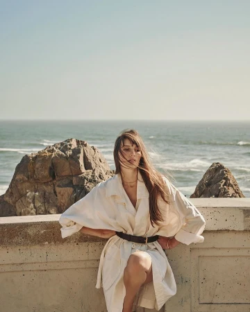 Woman in a white trench coat poses on a concrete wall near the ocean with rocks on the beach. Clear blue sky.