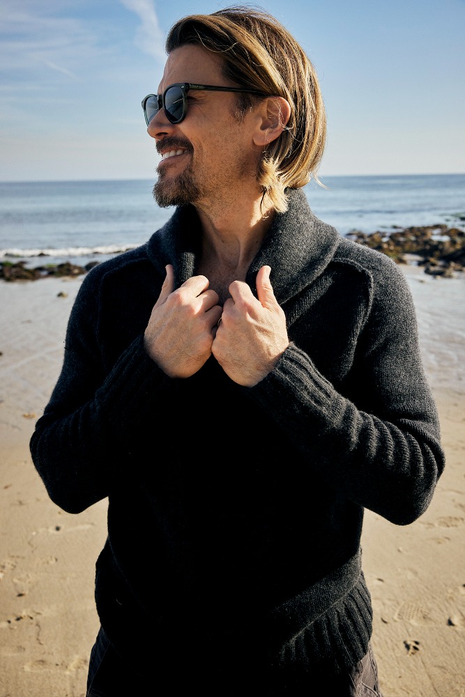 Man wearing dark sunglasses and a charcoal knit shawl-collar sweater standing on a sandy beach.