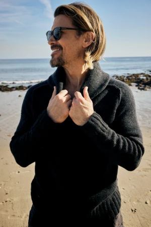 Man wearing dark sunglasses and a charcoal knit shawl-collar sweater standing on a sandy beach.