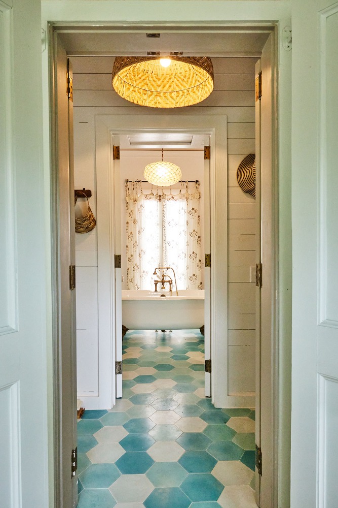 Indoor bathroom featuring turquoise geometric floor tiles, a freestanding white bathtub, and woven pendant lights visible through doorways.