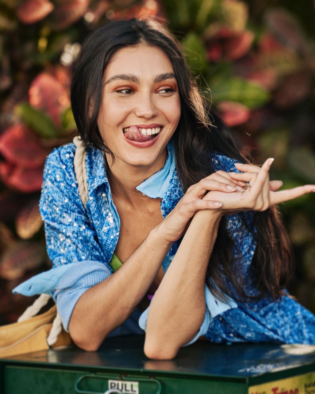 Portrait of a young woman with dark hair smiling and sticking out her tongue while leaning on a green utility box outdoors.