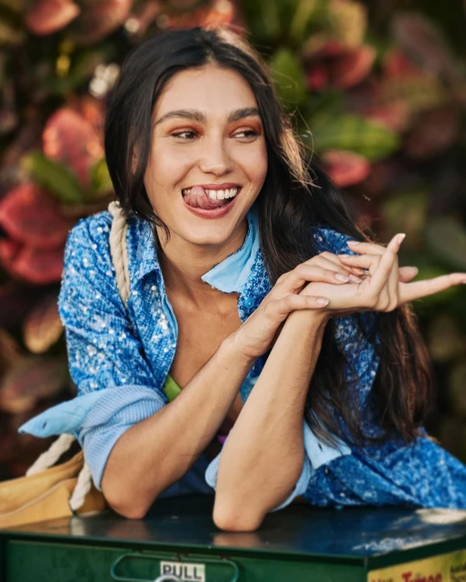 Portrait of a young woman with dark hair smiling and sticking out her tongue while leaning on a green utility box outdoors.
