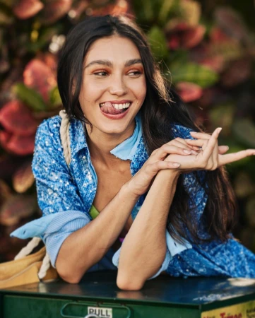 Portrait of a young woman with dark hair smiling and sticking out her tongue while leaning on a green utility box outdoors.