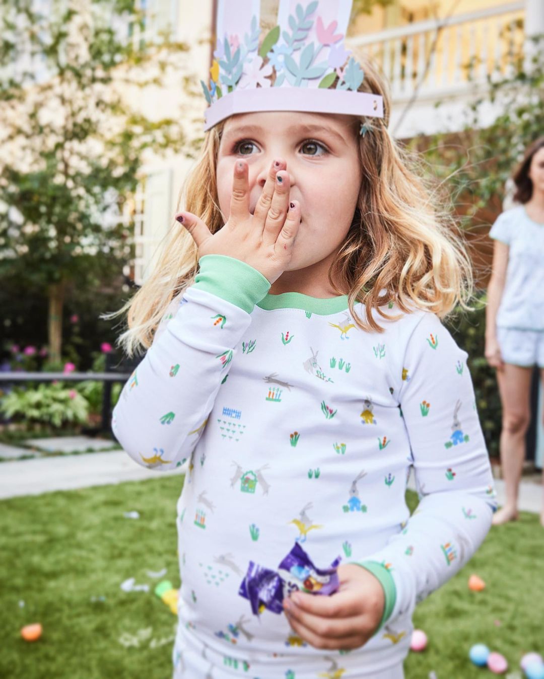 A young girl wearing a bunny ears headband stands on a green lawn holding a chocolate candy wrapper, with a woman in the background.