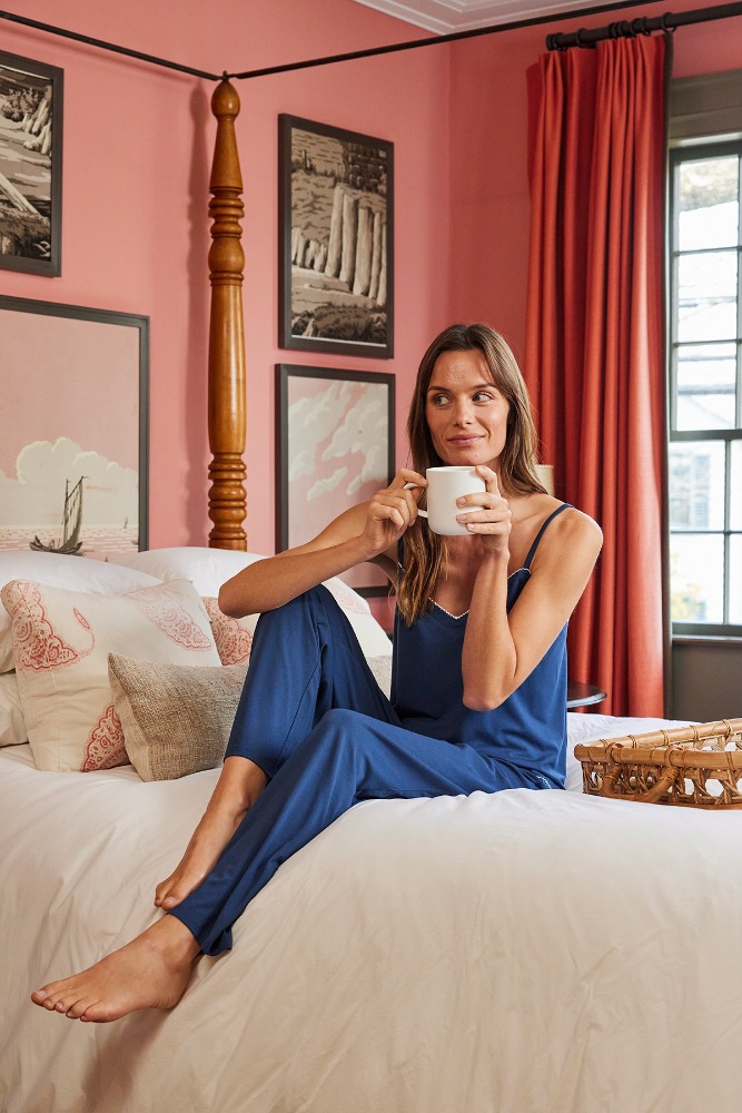 Brunette woman in blue pajamas sits on a white bed with patterned pillows, holding a white mug in a naturally lit room.