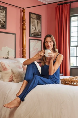 Brunette woman in blue pajamas sits on a white bed with patterned pillows, holding a white mug in a naturally lit room.