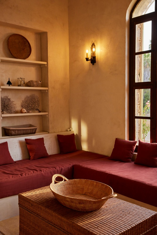 Living room interior with L-shaped built-in seating with red cushions, wooden coffee table, window, decorative wall shelf and sconce.