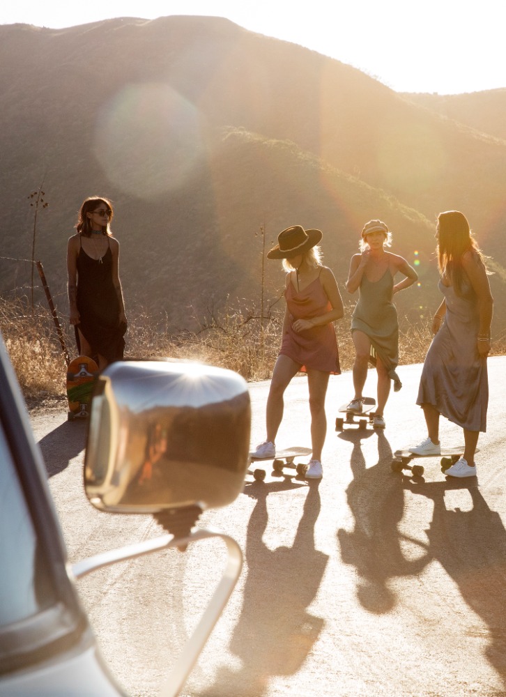 Four women in casual dresses pose on skateboards on a rural road, with a vintage car and mountain backdrop, at golden hour.