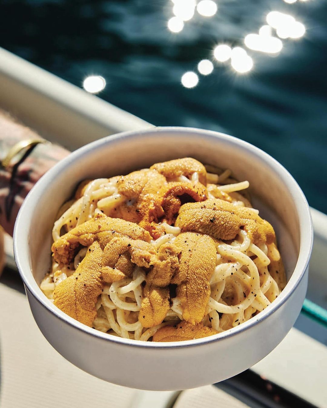 Overhead shot of a bowl of spaghetti topped with sea urchin roe, sitting on a boat with an ocean backdrop.