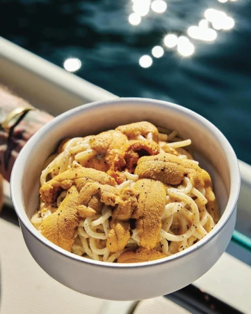 Overhead shot of a bowl of spaghetti topped with sea urchin roe, sitting on a boat with an ocean backdrop.