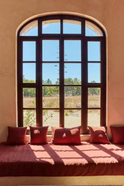 Window seat with rust-colored cushions and pillows in front of large window with view of field and trees on a sunny day.
