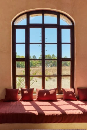 Window seat with rust-colored cushions and pillows in front of large window with view of field and trees on a sunny day.