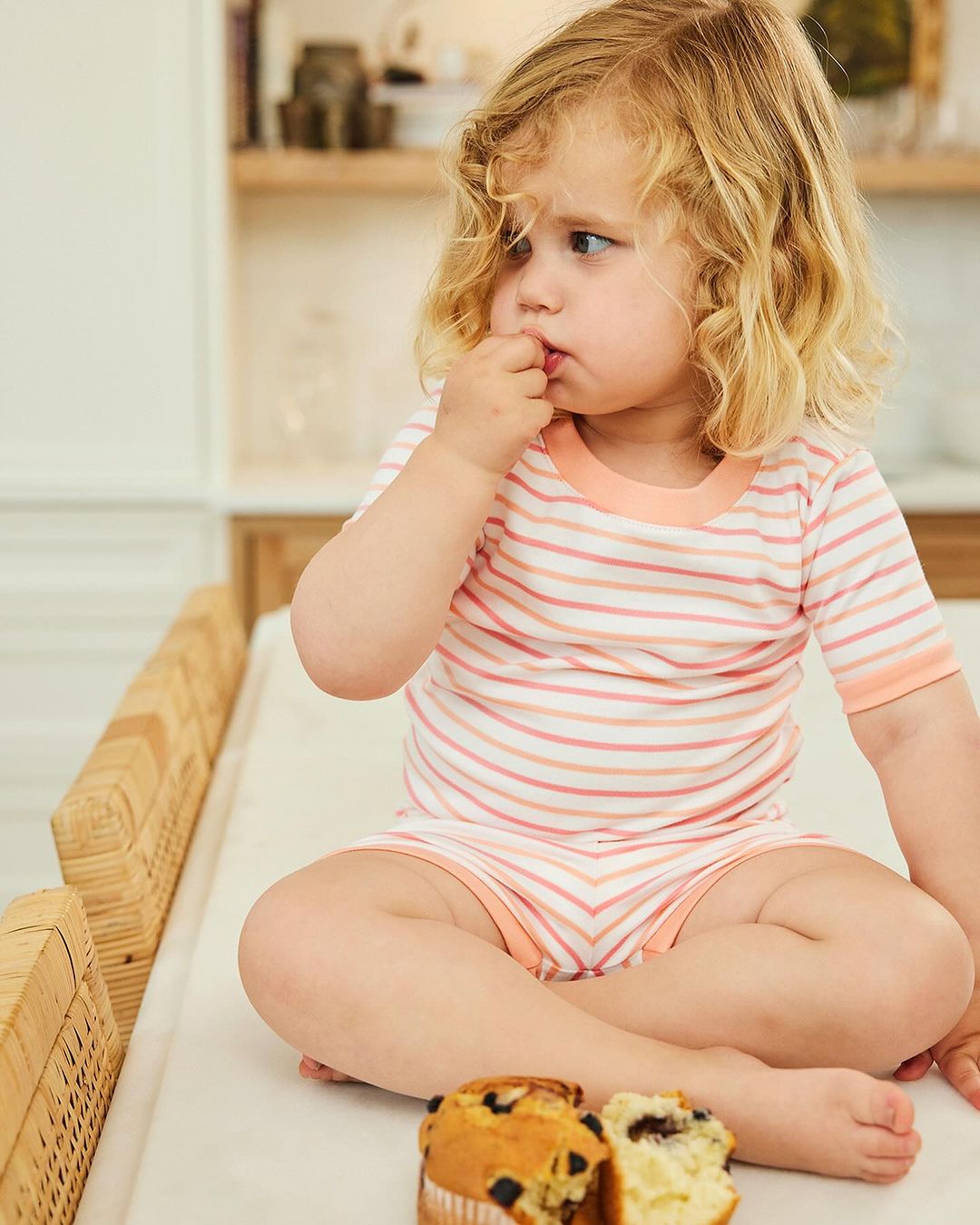 A young child with blonde curly hair sits on a wicker table, eating a muffin with berries in an indoor kitchen setting, shot in daylight.