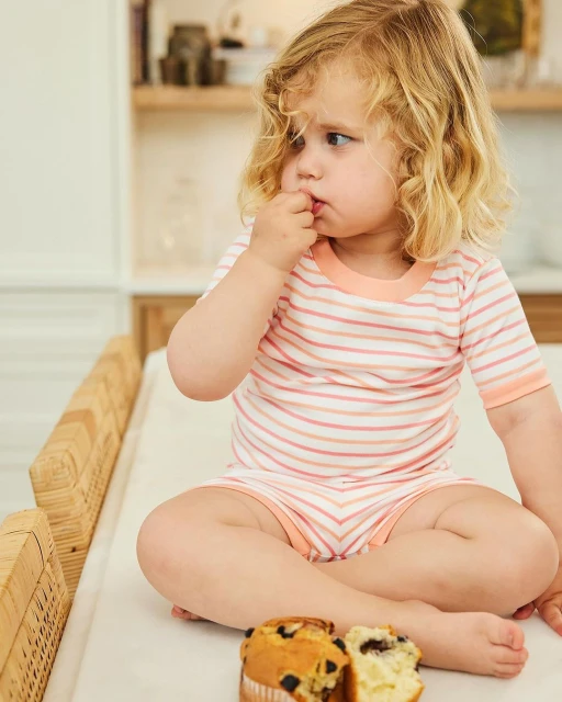 A young child with blonde curly hair sits on a wicker table, eating a muffin with berries in an indoor kitchen setting, shot in daylight.