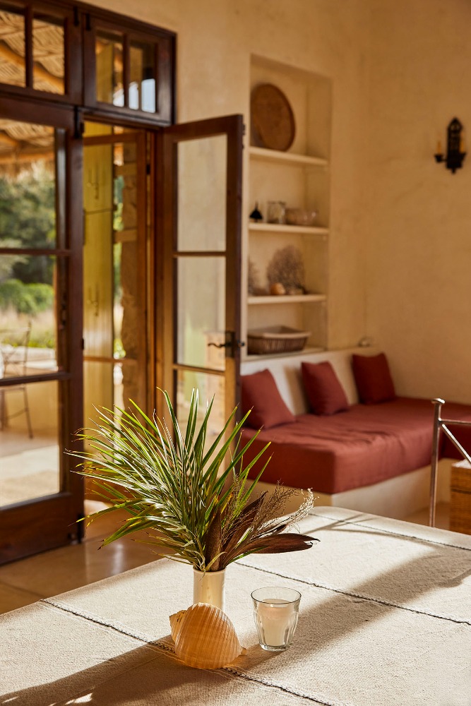 Indoor setting with a table displaying a shell, vase with greenery, and a glass, with a blurred view through open doors.