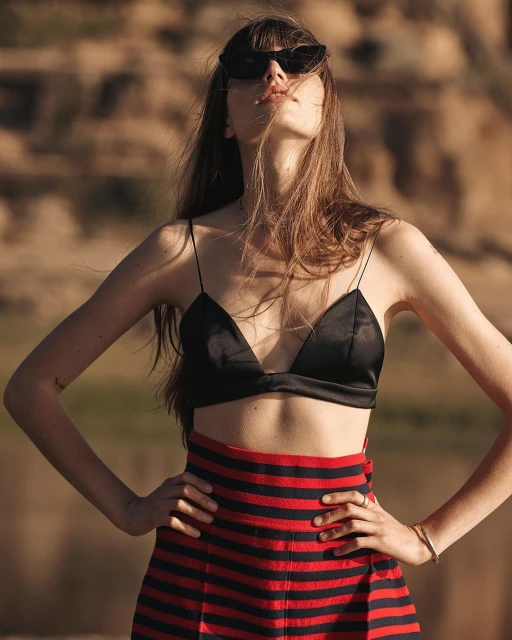 Woman in a black top and red striped skirt poses outdoors, wearing sunglasses with hands on hips against a blurred background.