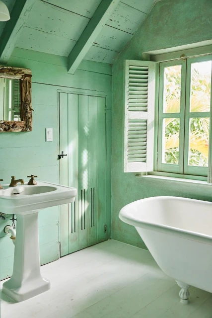 Bathroom interior with a pedestal sink, bathtub, and a shuttered window, all in shades of green and white, creating a serene atmosphere.