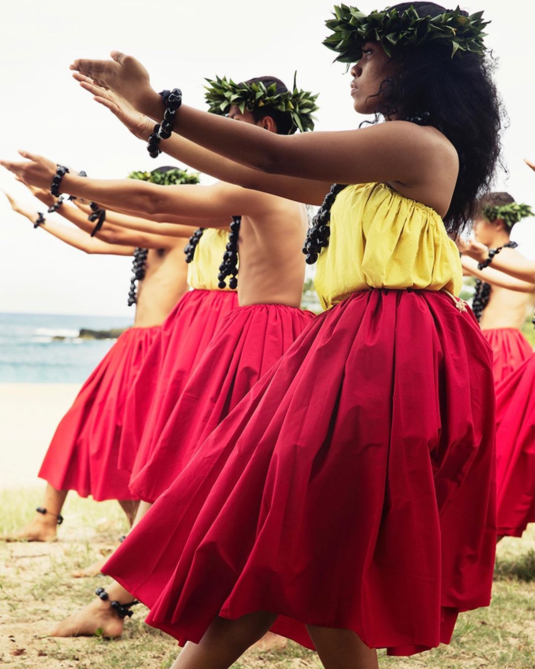 Group of women in hula skirts and leaf wreaths dance outdoors near the ocean, showcasing Hawaiian dance and culture with traditional attire.