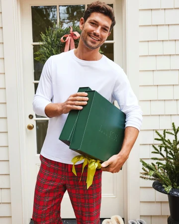 Man in pajamas smiles while holding a large green gift box with a yellow ribbon in front of a white shingle-clad door.