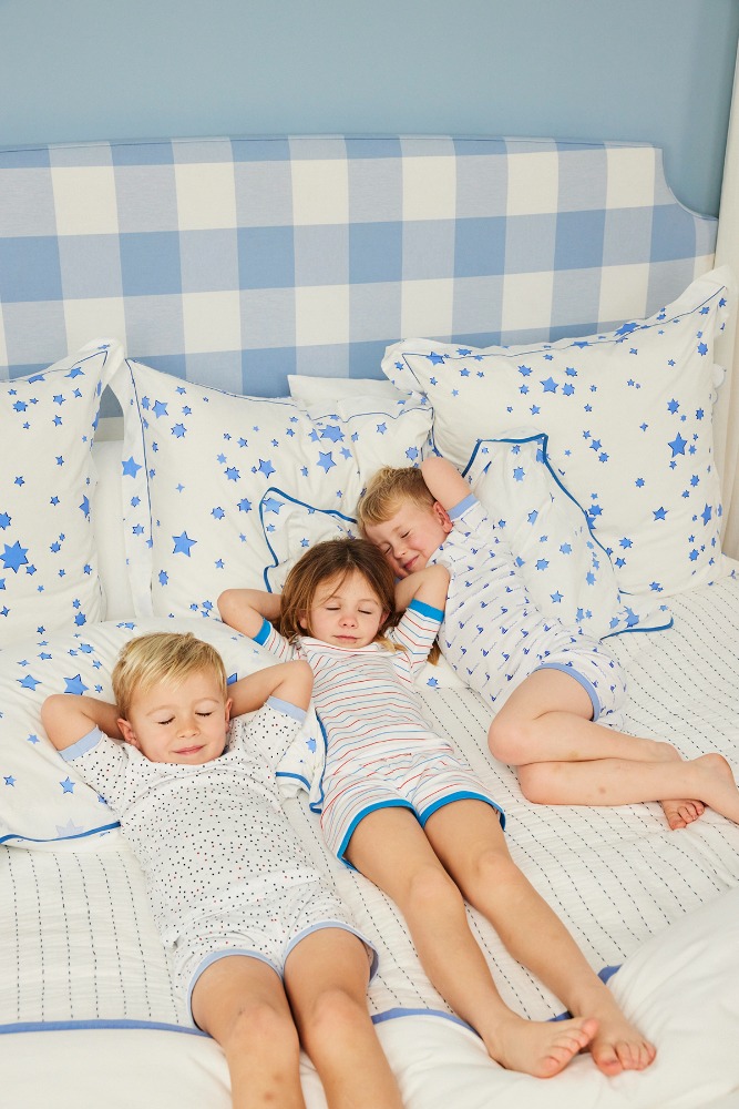 Three children, two boys and one girl, laying on their backs on a white bed with blue trim and pillow cases, with a blue and white buffalo check headboard.
