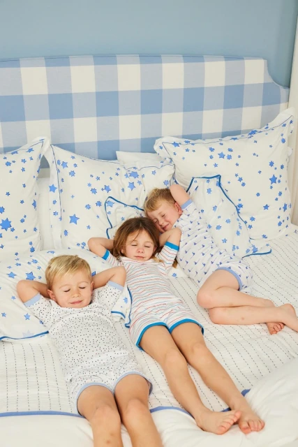 Three children, two boys and one girl, laying on their backs on a white bed with blue trim and pillow cases, with a blue and white buffalo check headboard.