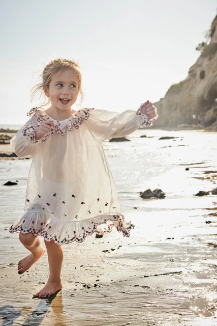 A young girl wearing a white dress runs barefoot on a sandy beach next to the ocean on a sunny day, with a cliff in the background.