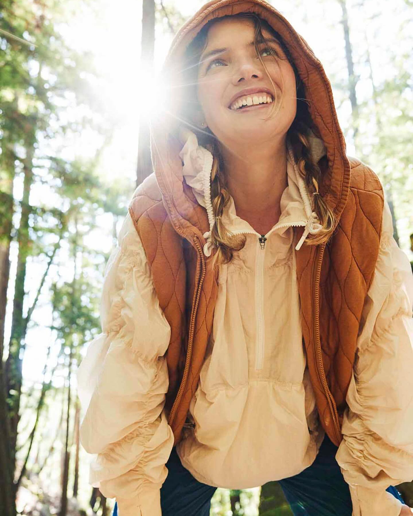 Woman in vest and puffer jacket looks up while standing among trees with sun flare in forest. Outdoor fashion photography.
