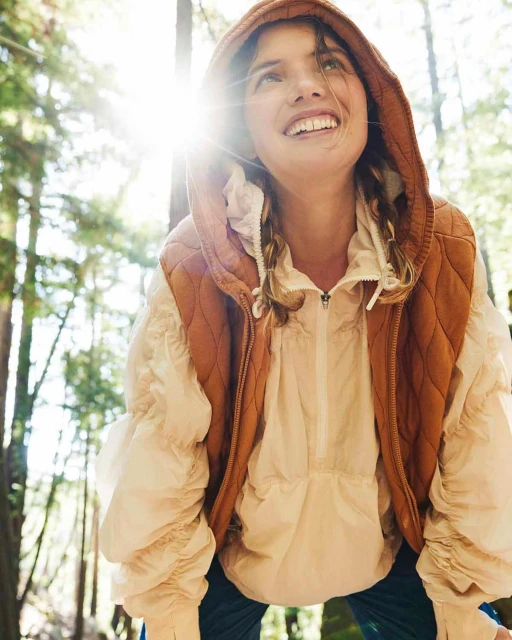 Woman in vest and puffer jacket looks up while standing among trees with sun flare in forest. Outdoor fashion photography.