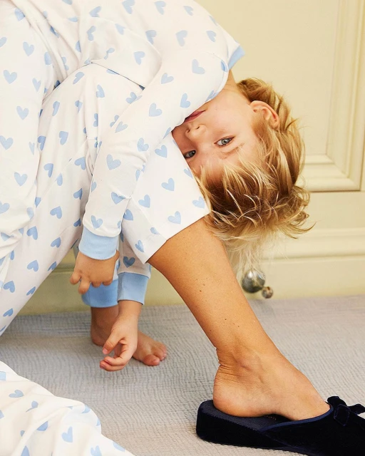 Child in heart-print pajamas rests head on adult leg indoors, both barefoot, with blue velvet slipper and muted background. 