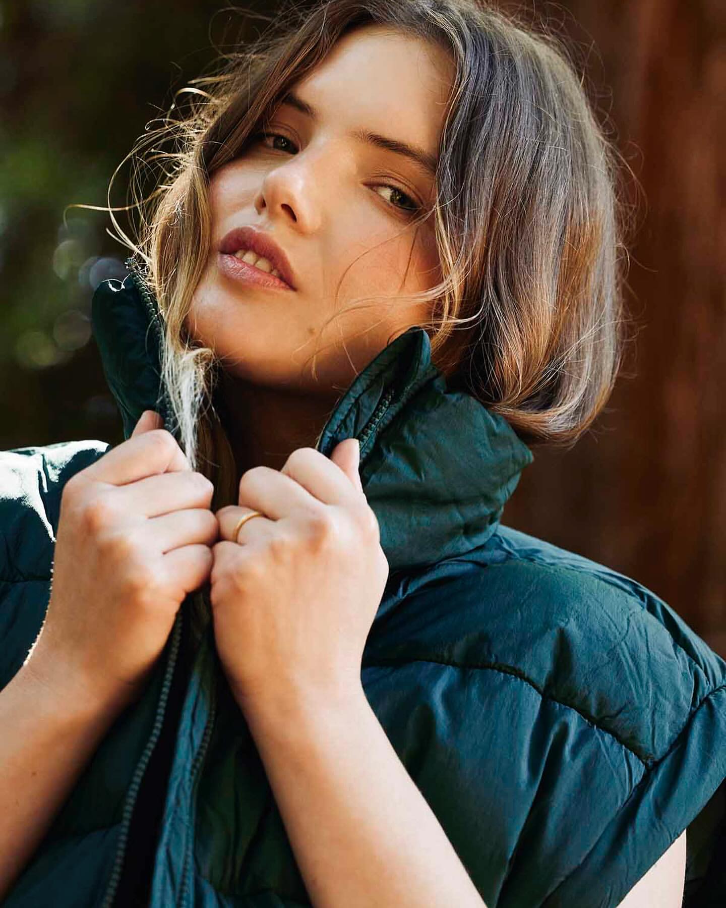 Portrait of a young woman with fair skin and light brown hair, wearing a puffy, dark teal vest, gazing at the camera against a natural backdrop.