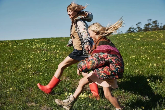 Two female models running across a grassy green meadow wearing patterned puffer jackets and tall red rain boots