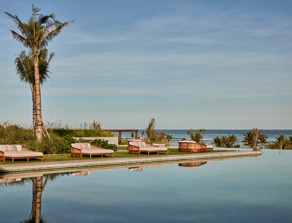 Resort pool terrace featuring lounge beds on green grass, palm trees, and ocean views under a clear sky.