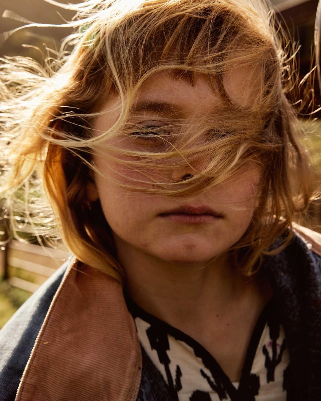 Close-up of a young person with light blonde hair partially covering their face, wearing a denim jacket and patterned shirt outdoors.