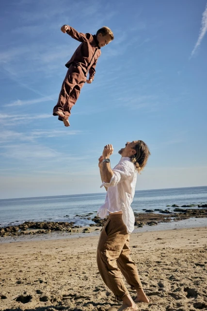 Father and son playfully interacting on a beach; the son is airborne, tossed by the father against a backdrop of blue sky and ocean.