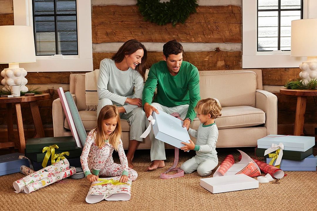 Family of four in matching pajamas sits on floor in living room, opening Christmas presents with wrapping paper and gift boxes.