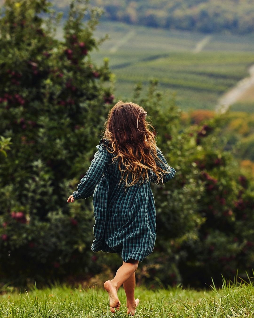 Barefoot child in green dress runs through lush field, back to camera, with orchards and rolling hills in background on a sunny day.