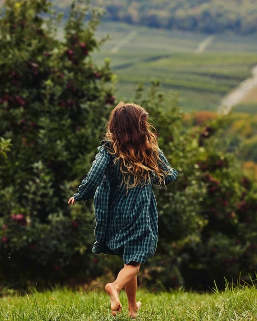 Barefoot child in green dress runs through lush field, back to camera, with orchards and rolling hills in background on a sunny day.